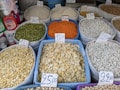 A variety of grains and legumes displayed in large sacks at a market, each labeled with a price tag. The sacks contain different types of beans, lentils, and seeds in various colors such as green, orange, white, and beige. The prices are written on cards placed on top of each sack. Nearby, there are bottles of detergent and paper towel rolls.