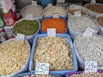 A variety of grains and legumes displayed in large sacks at a market, each labeled with a price tag. The sacks contain different types of beans, lentils, and seeds in various colors such as green, orange, white, and beige. The prices are written on cards placed on top of each sack. Nearby, there are bottles of detergent and paper towel rolls.