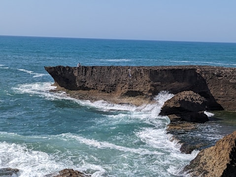 A rocky cliff juts out into a blue ocean with waves crashing against it. The sea is a vibrant turquoise near the shore, fading into a deep blue toward the horizon under a clear sky. A person stands on the cliff, adding a sense of scale to the natural landscape.