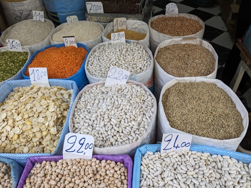 Various sacks filled with different types of grains and legumes are displayed at a marketplace. Each sack is labeled with a price tag indicating the cost per kilogram. The assortment includes lentils, beans, and seeds in a variety of colors and textures.