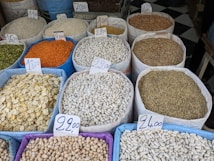 Various sacks filled with different types of grains and legumes are displayed at a marketplace. Each sack is labeled with a price tag indicating the cost per kilogram. The assortment includes lentils, beans, and seeds in a variety of colors and textures.