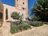 A garden with lush greenery and small flowering plants, surrounded by stone pathways. A tall, beige stone tower with small windows stands prominently to the left, accompanied by stone walls. Large leafy plants and palm trees are visible on the right, contributing to the tropical feel.