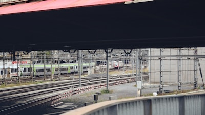 A train yard with multiple railway tracks and several passenger trains. The area is surrounded by industrial structures, with utility poles and wires crisscrossing above the tracks. The setting appears to be urban, with a sense of organization amidst the complex infrastructure.