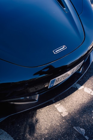 A close-up view of a dark-colored sports car's front end, highlighting the sleek design and shiny surface. The car features a distinct badge on the hood with a recognizable logo. The vehicle is parked on a textured asphalt surface with visible white markings.