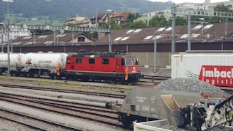 A red locomotive is positioned on railway tracks adjacent to several freight wagons. The scene includes tanker cars labeled 'jura cement' and a large container labeled 'imbach logistics.' Two individuals dressed in orange safety gear are standing near the train. In the foreground, there are heaps of gravel and industrial machinery. The backdrop features industrial buildings and a hilly landscape with greenery.