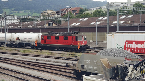 A red locomotive is positioned on railway tracks adjacent to several freight wagons. The scene includes tanker cars labeled 'jura cement' and a large container labeled 'imbach logistics.' Two individuals dressed in orange safety gear are standing near the train. In the foreground, there are heaps of gravel and industrial machinery. The backdrop features industrial buildings and a hilly landscape with greenery.