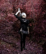 A person dressed in a black outfit and thigh-high boots stands in a forest with vivid red foliage. They are holding a sword over their shoulder, looking forward. The setting is mysterious and slightly fantastical.