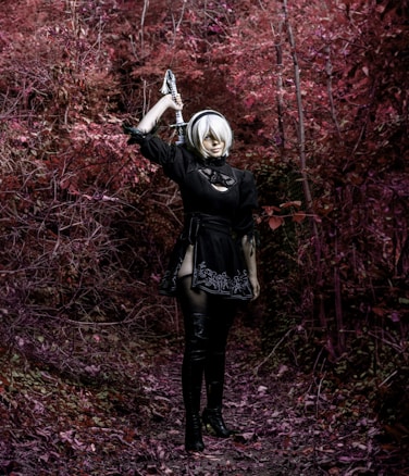 A person dressed in a black outfit and thigh-high boots stands in a forest with vivid red foliage. They are holding a sword over their shoulder, looking forward. The setting is mysterious and slightly fantastical.