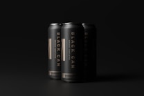 Three sleek, matte black cans are arranged with minimal lighting that highlights their metallic texture and bold typography. The lettering reads 'Black Can' and 'Mockup Templates' in a striking gold color, suggesting a premium or luxury product.