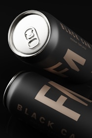 Two sleek black cans with a matte finish are placed at angles against a dark background. The label reads 'Black Can Mockup Templates' in minimalistic, modern typography. The top can is partially open, revealing a metallic pull tab.