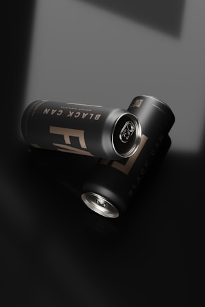 Two sleek, matte black cans with a minimalist design lie on a glossy black surface. The cans are labeled 'Black Can' in subtle lighting, enhancing the elegance of the presentation. The lighting creates soft reflections and shadows, adding to the modern aesthetic.