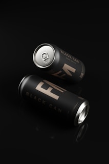 Two sleek matte black cans with metallic silver tops are placed against a dark reflective surface. The cans are labeled with the words 'BLACK CAN' in a stylish, minimalistic font. One can is upright while the other is slightly tilted.