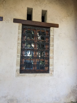 A rustic window set in a stone wall, featuring decorative wrought iron bars. The wall is textured and appears weathered, suggesting an old building. The wooden window frame is visible behind the intricate black metalwork.