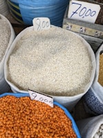 Various sacks of grains and legumes are displayed with handwritten price tags. The sacks contain what appear to be sesame seeds, lentils, and other grains. The price tags show amounts written in a currency notation. The image likely depicts a market or store setting.