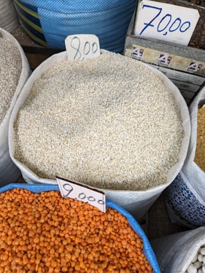 Various sacks of grains and legumes are displayed with handwritten price tags. The sacks contain what appear to be sesame seeds, lentils, and other grains. The price tags show amounts written in a currency notation. The image likely depicts a market or store setting.