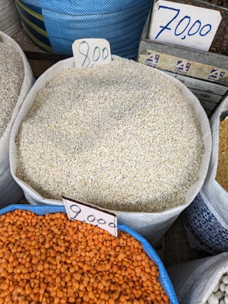 Various sacks of grains and legumes are displayed with handwritten price tags. The sacks contain what appear to be sesame seeds, lentils, and other grains. The price tags show amounts written in a currency notation. The image likely depicts a market or store setting.