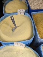 Several large blue bags filled with yellow grains or semolina are displayed in a market setting. A metal scoop is partially buried in one of the bags, indicating the product is sold loose by weight. Handwritten price tags are placed on each bag.