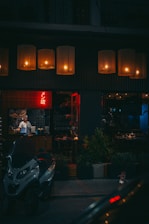 A dimly lit street scene features a cozy, warmly illuminated restaurant with large hanging lamps across the entrance. A chef or waiter is visible inside through the window, standing near a counter. Outside, a parked motorcycle is in the foreground, and potted plants line the sidewalk.