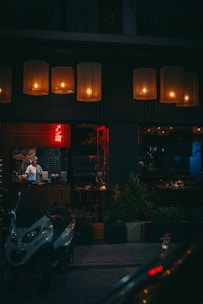 A dimly lit street scene features a cozy, warmly illuminated restaurant with large hanging lamps across the entrance. A chef or waiter is visible inside through the window, standing near a counter. Outside, a parked motorcycle is in the foreground, and potted plants line the sidewalk.