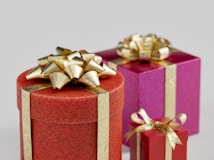 Three brightly colored gift boxes are adorned with shiny golden ribbons. The largest gift is round and red with a speckled texture, topped with an elaborate gold bow. Behind it, a medium-sized magenta square box also has a gold ribbon and bow. The smallest box is red, featuring a simple gold ribbon bow.