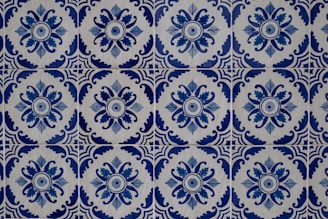 Intricate blue and white ceramic tiles featuring repetitive floral and geometric patterns. The design showcases a circular floral motif surrounded by ornate, symmetrical shapes, creating a harmonious and classic aesthetic.