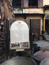 A sign featuring text in Arabic is placed in front of a historic wooden doorway. The surroundings include a large tree and multiple cooking pots. Above the sign, a building facade displays colorful signage advertising food and drinks, with open windows revealing a glimpse of the interior.