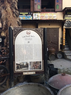 A sign featuring text in Arabic is placed in front of a historic wooden doorway. The surroundings include a large tree and multiple cooking pots. Above the sign, a building facade displays colorful signage advertising food and drinks, with open windows revealing a glimpse of the interior.