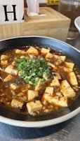 A bowl filled with a spicy dish featuring cubed tofu submerged in a rich, reddish-brown sauce, garnished with chopped green onions. The dish is set on a metallic table, and there are wooden and glass elements visible in the background.