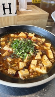 A bowl filled with a spicy dish featuring cubed tofu submerged in a rich, reddish-brown sauce, garnished with chopped green onions. The dish is set on a metallic table, and there are wooden and glass elements visible in the background.