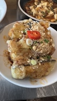 A dish with battered and fried pieces of eggplant, garnished with chopped green onions, peanuts, sesame seeds, and a slice of red chili. In the background, there is another dish that appears to be tofu in a rich sauce with green onions and possibly other vegetables.