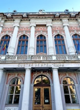 A neoclassical building facade with tall columns and ornate architectural details. The structure features multiple arched windows with decorative embellishments, such as sculpted figures and floral motifs. Above the entrance, the words 'Curtea de Apel Cluj' are inscribed. Two flags are displayed, one on each side of the building.