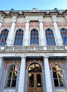 A neoclassical building facade with tall columns and ornate architectural details. The structure features multiple arched windows with decorative embellishments, such as sculpted figures and floral motifs. Above the entrance, the words 'Curtea de Apel Cluj' are inscribed. Two flags are displayed, one on each side of the building.