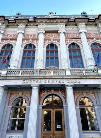 A neoclassical building facade with tall columns and ornate architectural details. The structure features multiple arched windows with decorative embellishments, such as sculpted figures and floral motifs. Above the entrance, the words 'Curtea de Apel Cluj' are inscribed. Two flags are displayed, one on each side of the building.