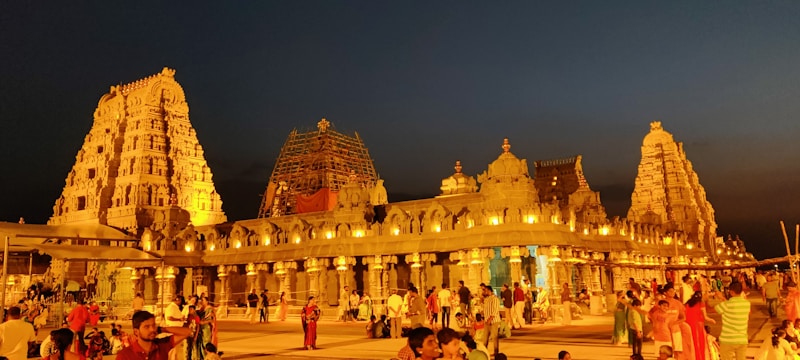 A vibrant and bustling scene at a grand temple complex during the evening, with the temple beautifully illuminated against a dark sky. The architecture is intricate and ornate, featuring tall towers and detailed carvings, with scaffolding visible on one tower. A large crowd of people, many dressed in colorful traditional attire, are gathered in the foreground, contributing to a lively and festive atmosphere.