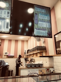 A cafe area with a striped pink and white wall in the background. Two employees are working behind the counter; one is seen wearing an apron and preparing an item. Overhead, there are menu boards displaying various drink options such as cold coffee, smoothies, iced beverages, and hot beverages. Shelves with glasses and cups are mounted on the wall, and safety signage is visible near the counter.