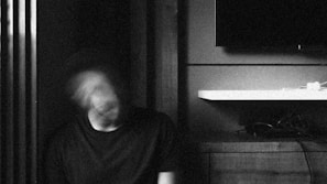 A blurred figure sits with their head in motion, creating a ghostly effect. They are positioned against a backdrop of vertical slats and a wall-mounted shelf or surface with a television nearby. The setting seems to be indoors, with a monochrome and grainy texture.