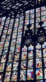 A series of vibrant stained glass windows depicting various historical and religious scenes. Each panel is intricately designed, showcasing figures in dynamic poses and a range of expressions. The artwork is richly detailed, with elements such as robes, facial features, and backgrounds meticulously crafted.