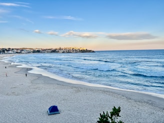 A sandy beach stretches along the coastline with gentle ocean waves approaching the shore. Scattered individuals are walking along the beach, and a blue tent is set up on the sand. In the distance, a cluster of buildings is visible under a clear, blue sky with a few clouds.