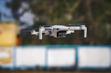 A compact drone in mid-flight with a camera mounted on the front. It features four propellers, each attached to an arm extending from the body. The background consists of blurred colors, indicating shallow depth of field and motion blur.