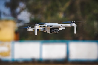 A compact drone in mid-flight with a camera mounted on the front. It features four propellers, each attached to an arm extending from the body. The background consists of blurred colors, indicating shallow depth of field and motion blur.
