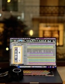 A laptop displaying an audio editing or digital audio workstation (DAW) software with various tracks and controls on the screen. A pair of headphones rests beside the laptop, suggesting an audio production or mixing environment. The blurred background indicates an indoor setting, possibly near a window, with warm lighting.