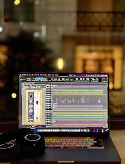 A laptop displaying an audio editing or digital audio workstation (DAW) software with various tracks and controls on the screen. A pair of headphones rests beside the laptop, suggesting an audio production or mixing environment. The blurred background indicates an indoor setting, possibly near a window, with warm lighting.