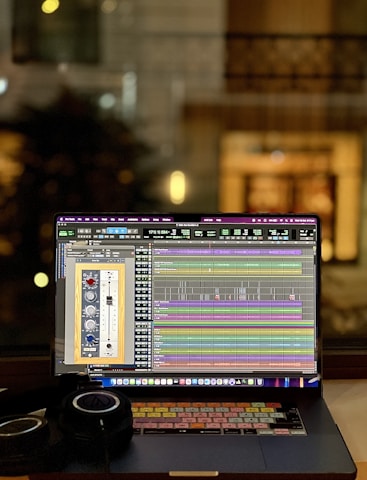 A laptop displaying an audio editing or digital audio workstation (DAW) software with various tracks and controls on the screen. A pair of headphones rests beside the laptop, suggesting an audio production or mixing environment. The blurred background indicates an indoor setting, possibly near a window, with warm lighting.