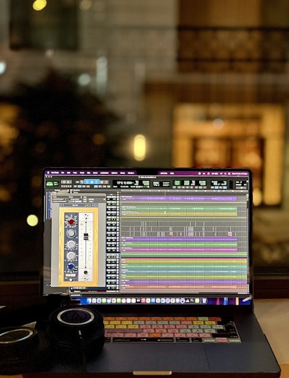 A laptop displaying an audio editing or digital audio workstation (DAW) software with various tracks and controls on the screen. A pair of headphones rests beside the laptop, suggesting an audio production or mixing environment. The blurred background indicates an indoor setting, possibly near a window, with warm lighting.