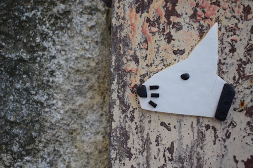 A creative depiction of a dog's head is crafted with white material and black accents for the nose, eye, and whiskers. The artwork is affixed to a weathered wall with peeling paint that features a mix of gray, beige, and reddish-brown colors.