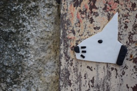 A creative depiction of a dog's head is crafted with white material and black accents for the nose, eye, and whiskers. The artwork is affixed to a weathered wall with peeling paint that features a mix of gray, beige, and reddish-brown colors.