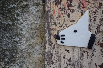 A creative depiction of a dog's head is crafted with white material and black accents for the nose, eye, and whiskers. The artwork is affixed to a weathered wall with peeling paint that features a mix of gray, beige, and reddish-brown colors.