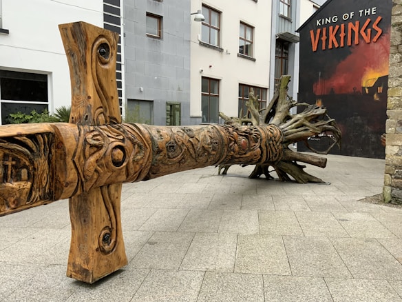 A large, intricately carved wooden sculpture resembling a tree lays horizontally on a paved courtyard. In the background, there is a mural with bold lettering that says 'King of the Vikings' set against a dark, smoky backdrop. The sculpture features detailed patterns and faces carved into it.