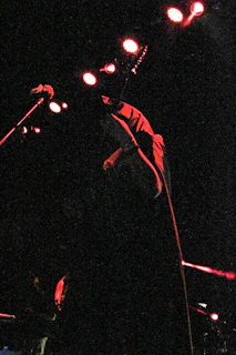 A shadowy figure with dark makeup and gothic attire playing an electric guitar under dim purple stage lights.