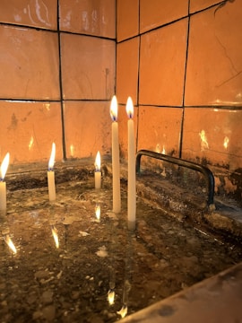 Several lit candles are placed in what appears to be a shallow basin or tray filled with water. The candles are burning steadily, and their reflections are visible in the water below. The background consists of uneven, orange-brown tiled walls with some visible wear and marks.
